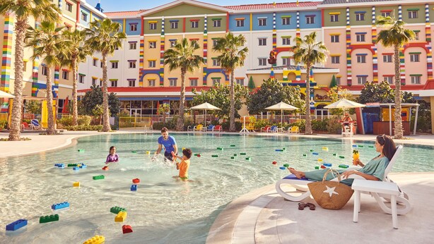 Une zone de piscine d’hôtel aux couleurs vives, avec une eau cristalline, des palmiers et un bâtiment à la façade rayée.