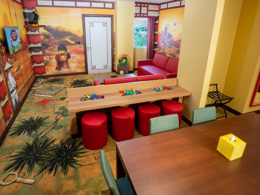 A room with a wooden table, red stools, a sofa, and a wall with a mural.