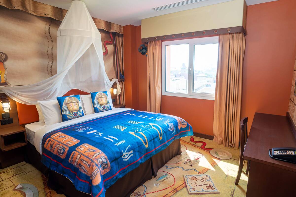 Una habitación de hotel con cama, escritorio, ordenador, ventana y cortina.