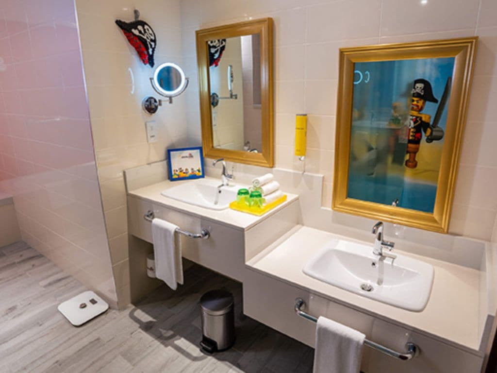 Un baño con dos lavamanos, un espejo, una báscula y un cubo de basura.