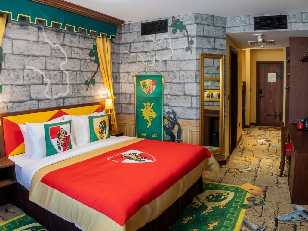 Quarto de hotel com parede de tijolos, cama com colcha vermelha e faixa verde e dourada.