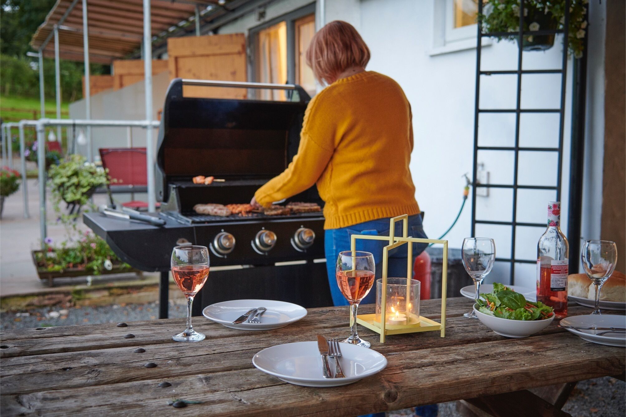 Uma pessoa fazendo churrasco ao ar livre, com uma mesa posta para refeição, incluindo pratos, talheres e copos.