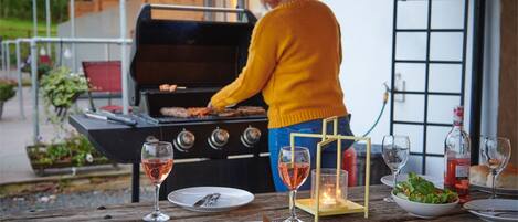 Uma pessoa fazendo churrasco ao ar livre, com uma mesa posta para refeição, incluindo pratos, talheres e copos.