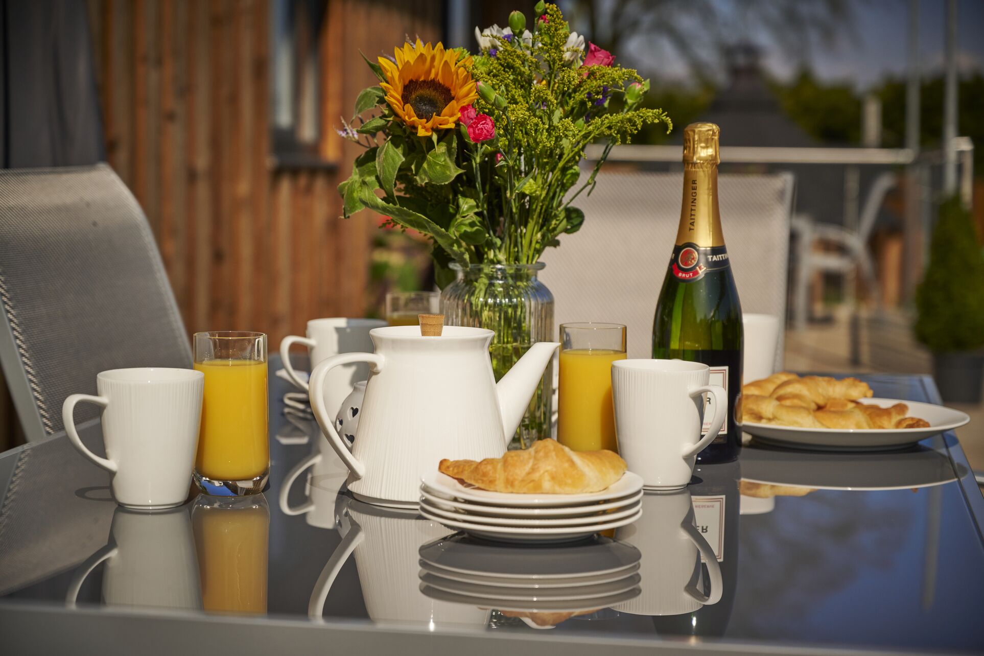 A breakfast table with a coffee pot, cups, a glass of orange juice, a bottle of champagne, a plate of croissants, and a vase of flowers.