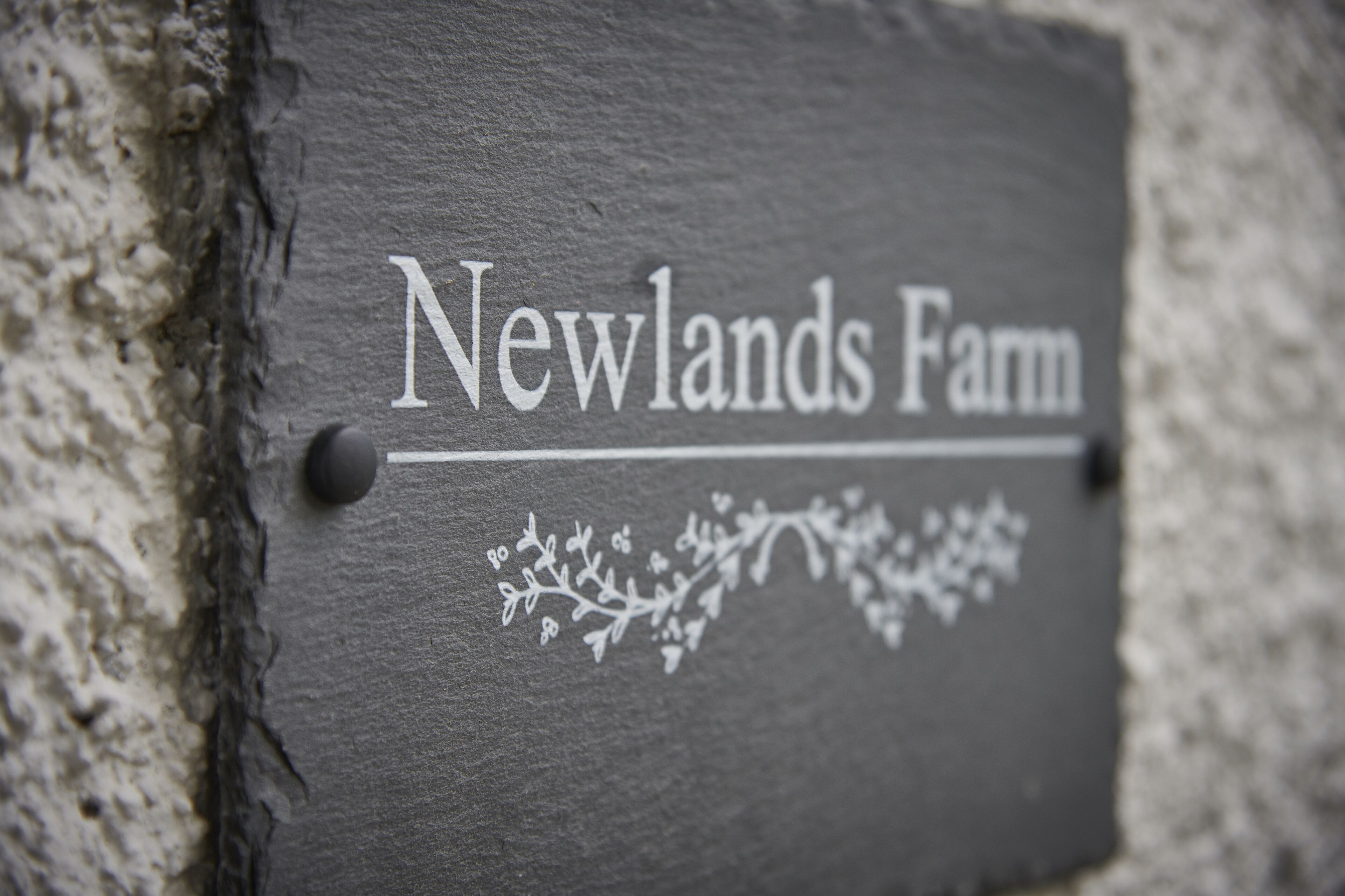 Egy palából készült tábla, amelyre rá van gravírozva a „Newlands Farm” felirat, és virágmintával díszített.