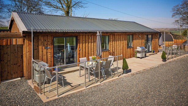 A wooden building with a metal roof, a patio with outdoor seating, and a small garden area. - Newlands Farm Stables (Kendal)