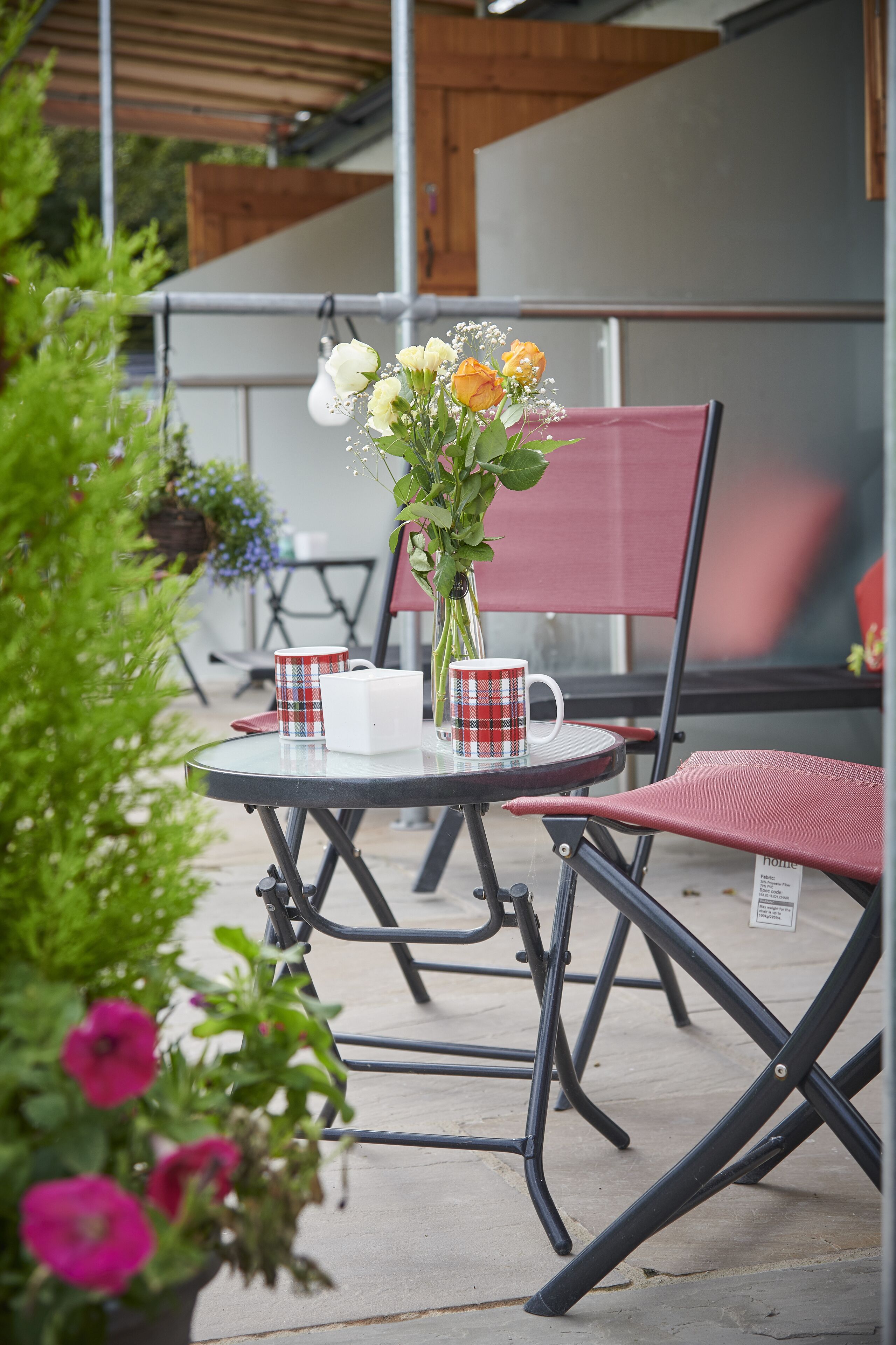 Un espacio al aire libre con una mesa redonda, dos sillas rojas y una planta en maceta con flores.