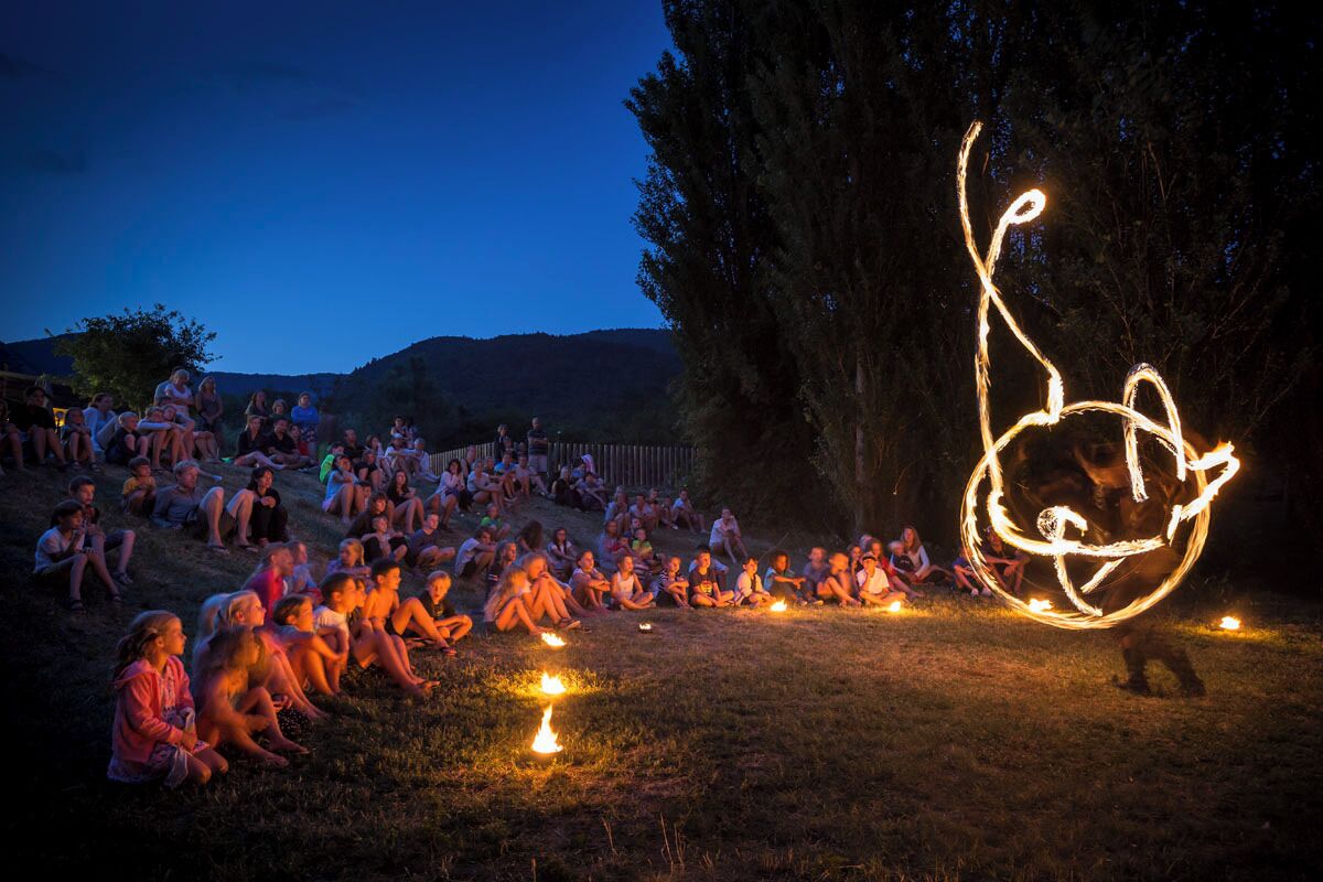 A group of people sitting on grass at night, with a fire performer creating a large, illuminated design in the air.