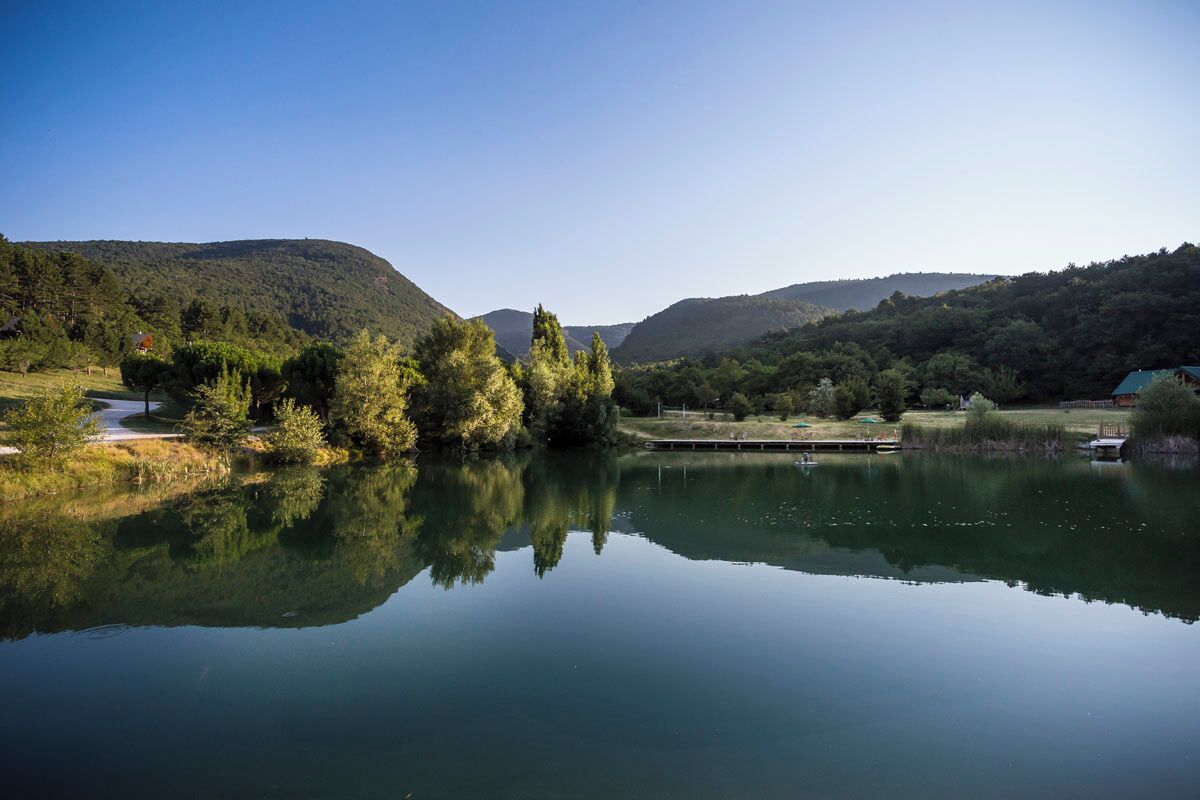 Un lago tranquilo con reflejos de árboles y colinas, rodeado de vegetación y algunos edificios.