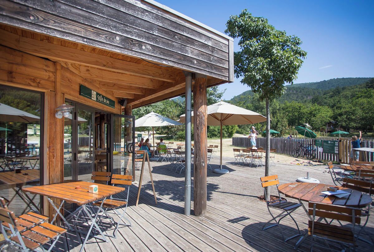 Terraza de madera al aire libre con mesas y sillas, rodeada de sombrillas y vistas a una ladera boscosa.