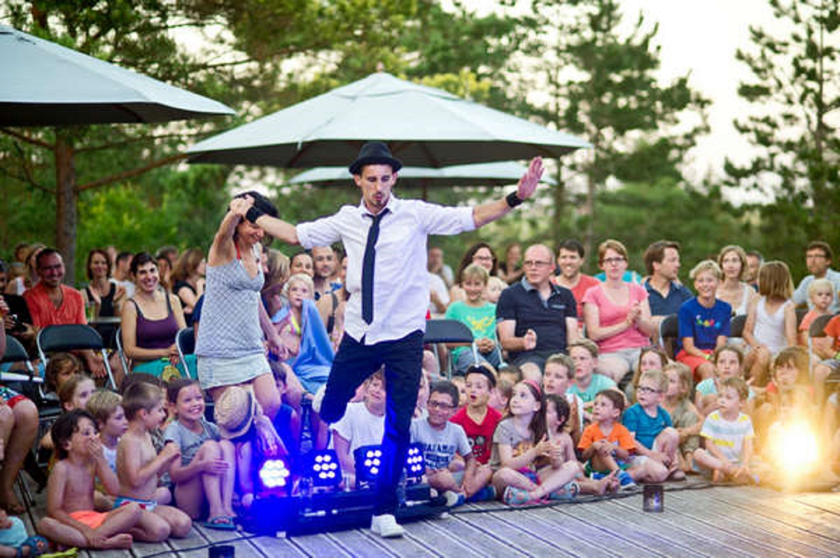 A stage with a performer in a white shirt and black tie, an audience seated on the ground, and a large umbrella in the background.