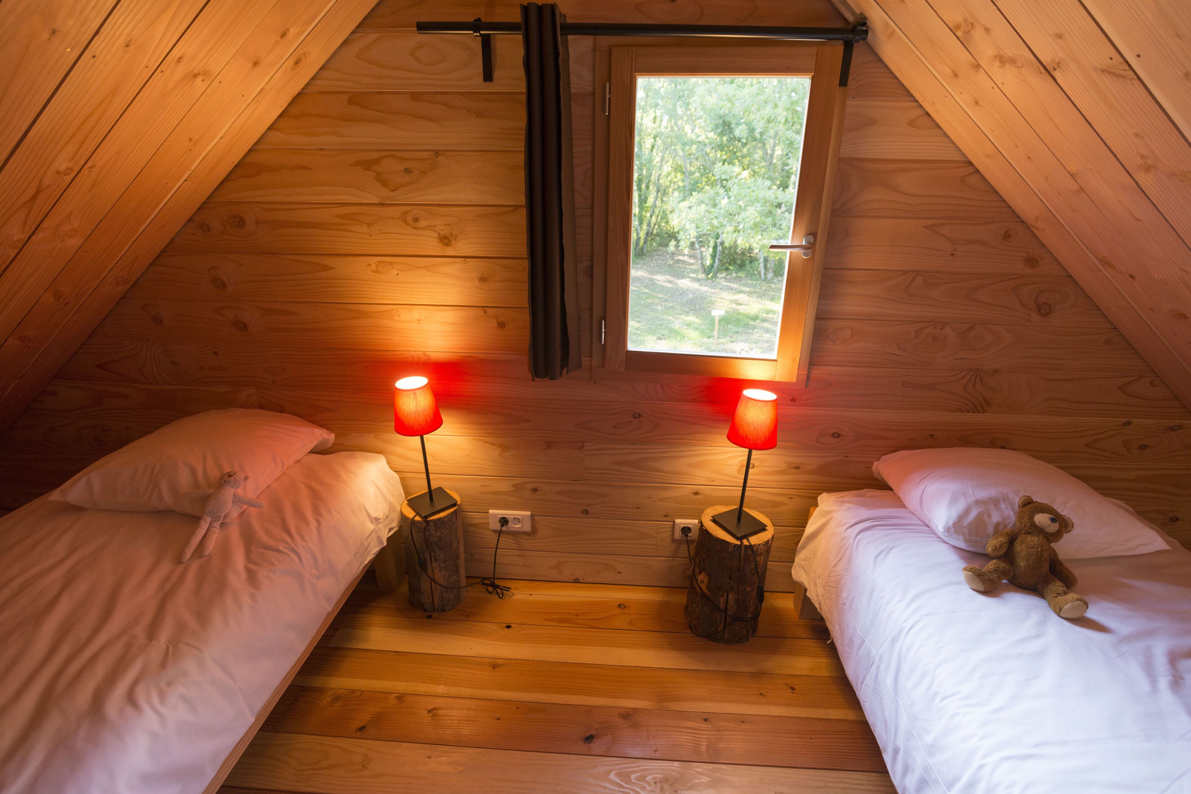 A cozy attic bedroom with two beds, each with a wooden nightstand and a lamp. A window provides a view of a forested area.