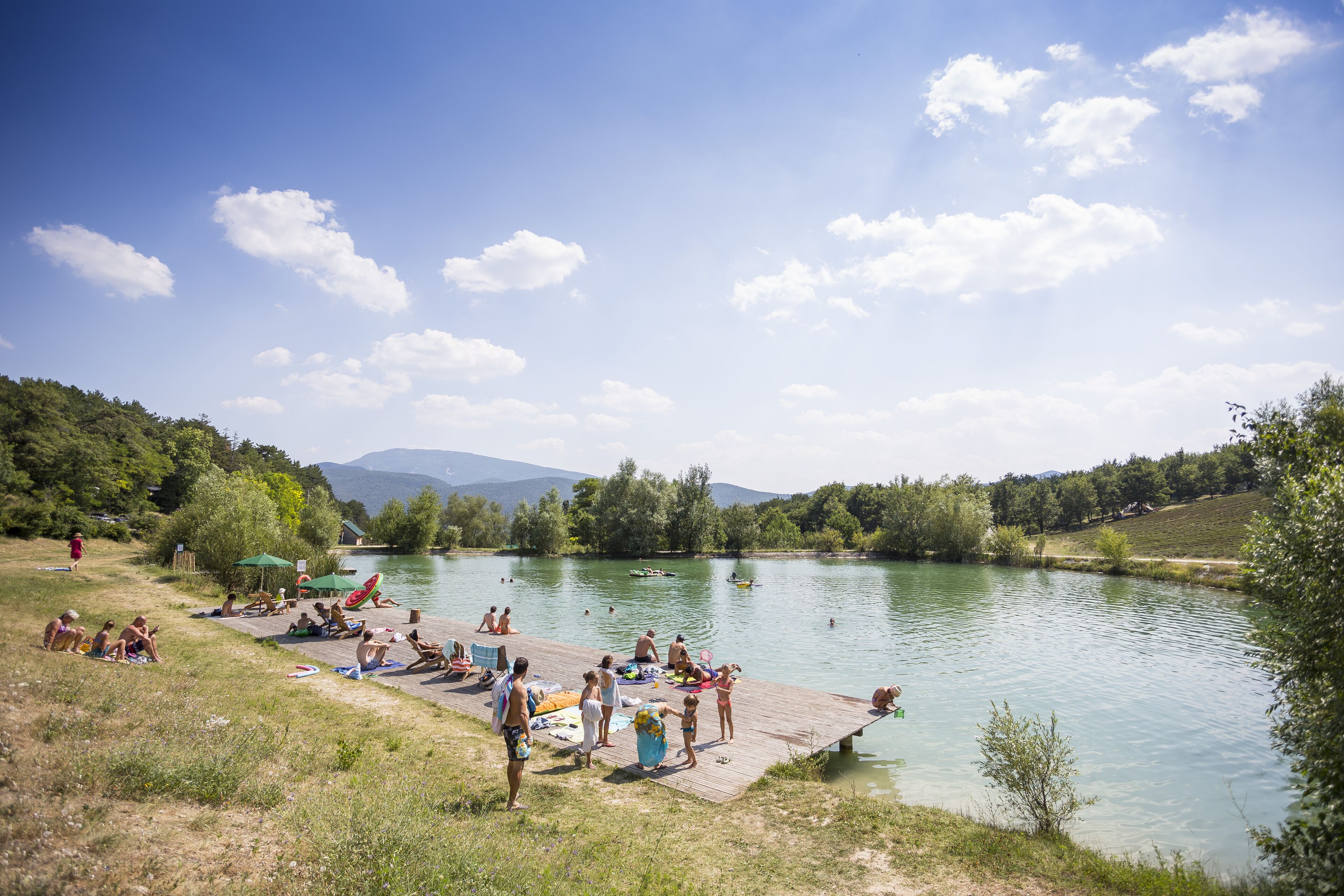 A lakeside scene with people relaxing by the water, a dock, and greenery in the background.