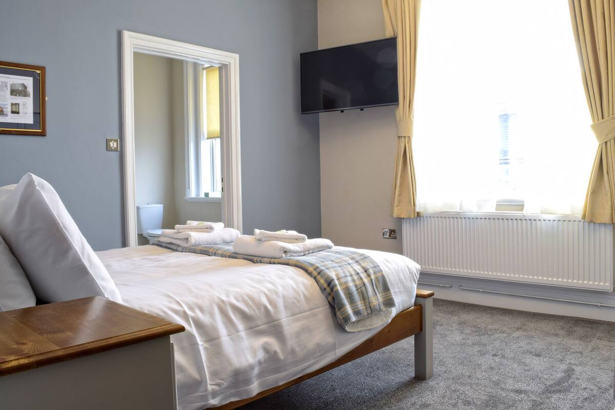 Hotellihuone, jossa on sänky, yöpöytä, taulutelevisio, ikkuna verhoilla ja patteri.