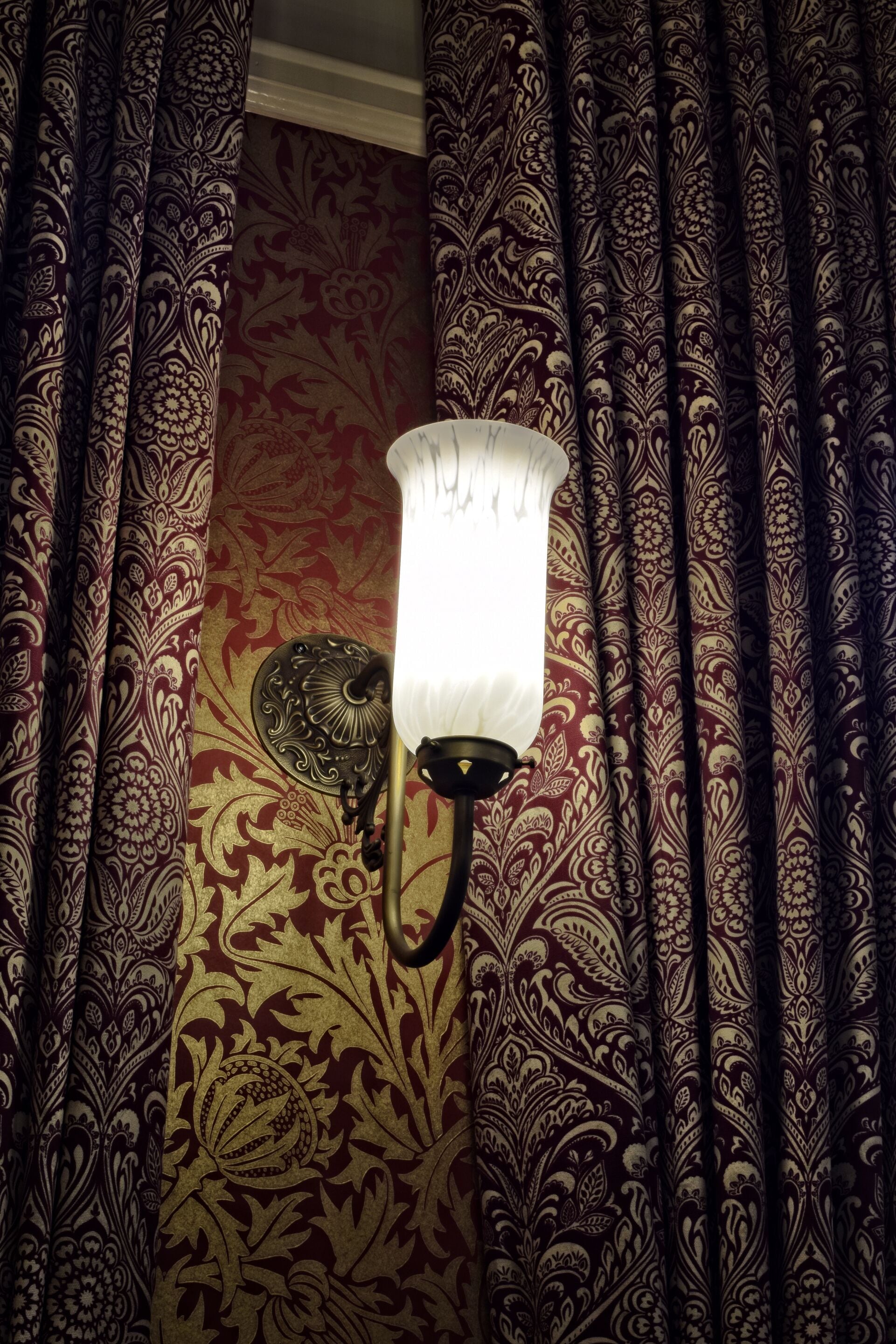 A wall-mounted lamp with a white shade, set against a patterned wallpaper with floral designs.