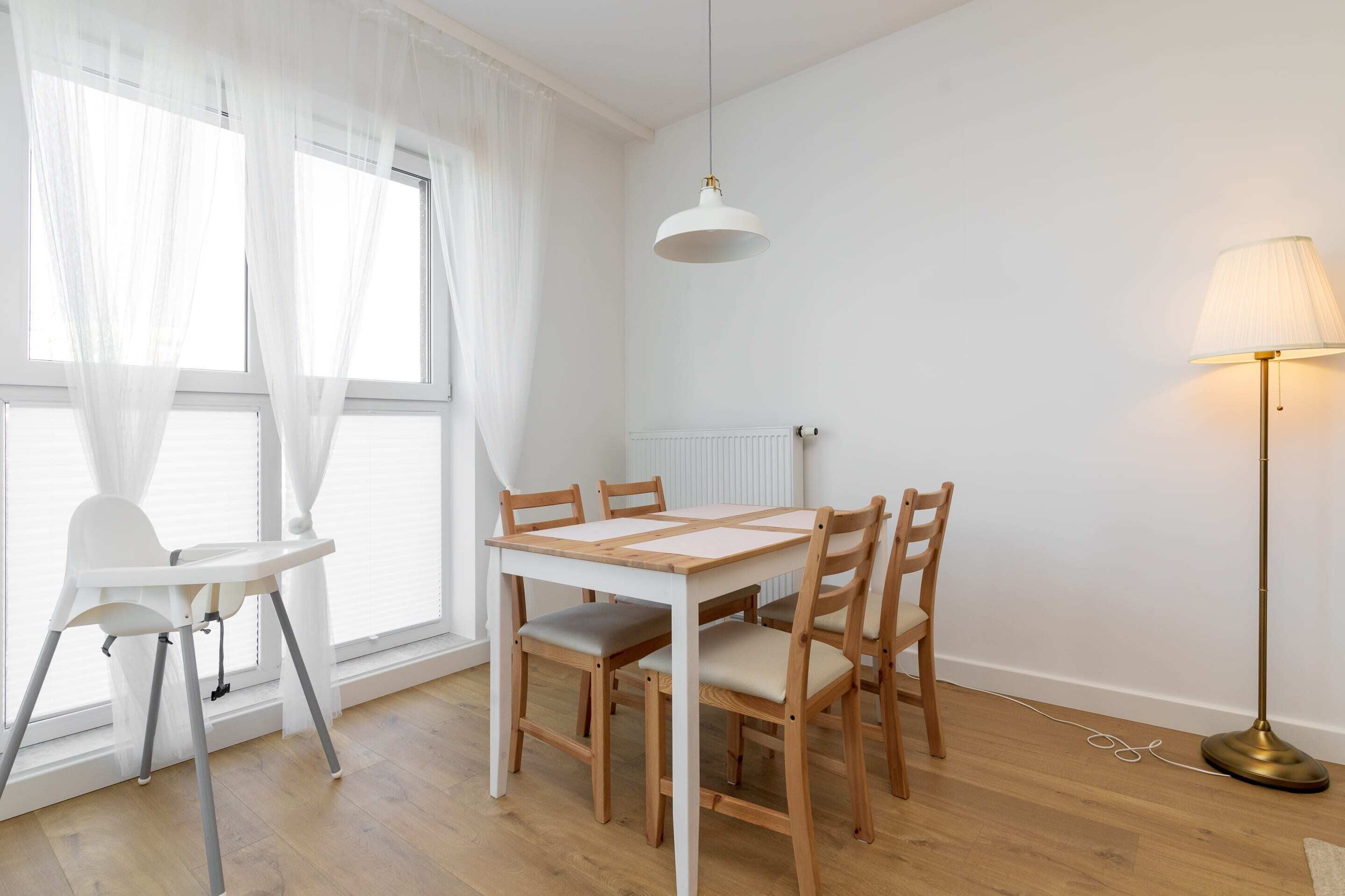 Un comedor con mesa y sillas blancas, una lámpara de pie y cortinas de gasa.