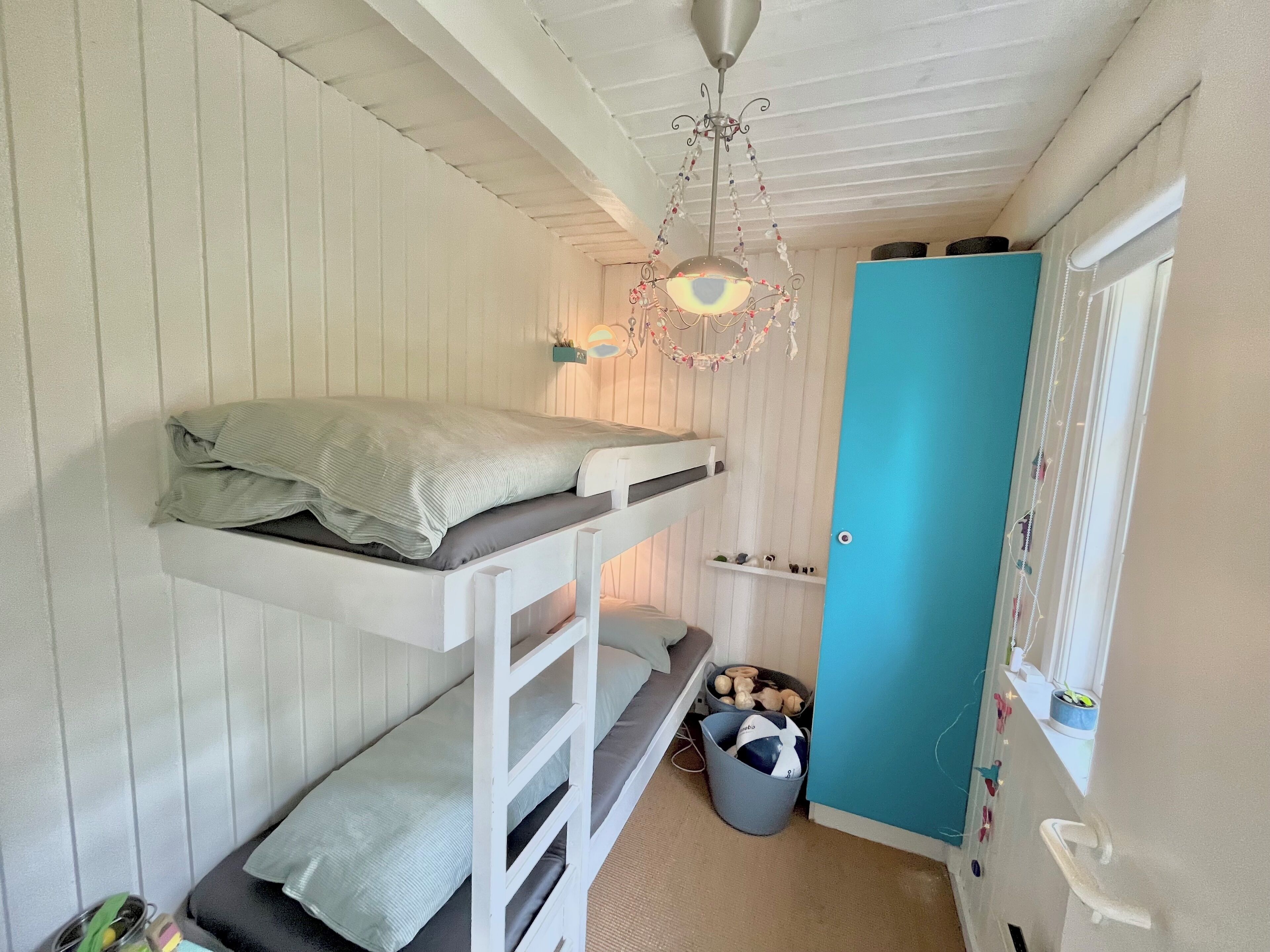 Een kleine kamer met een stapelbed, een klein bankje, een plafondlamp en een deur.