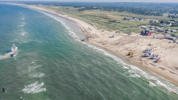 Vue aérienne d’une zone côtière avec une plage, des bateaux et un petit village composé de maisons et d’immeubles.