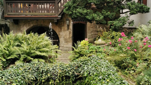 Un edificio de piedra con balcón, una pequeña fuente de agua y abundante vegetación.