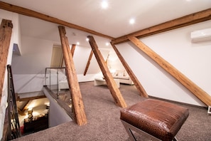 An attic room with exposed wooden beams, a brown leather ottoman, a white sofa, and a glass railing overlooking the lower level.