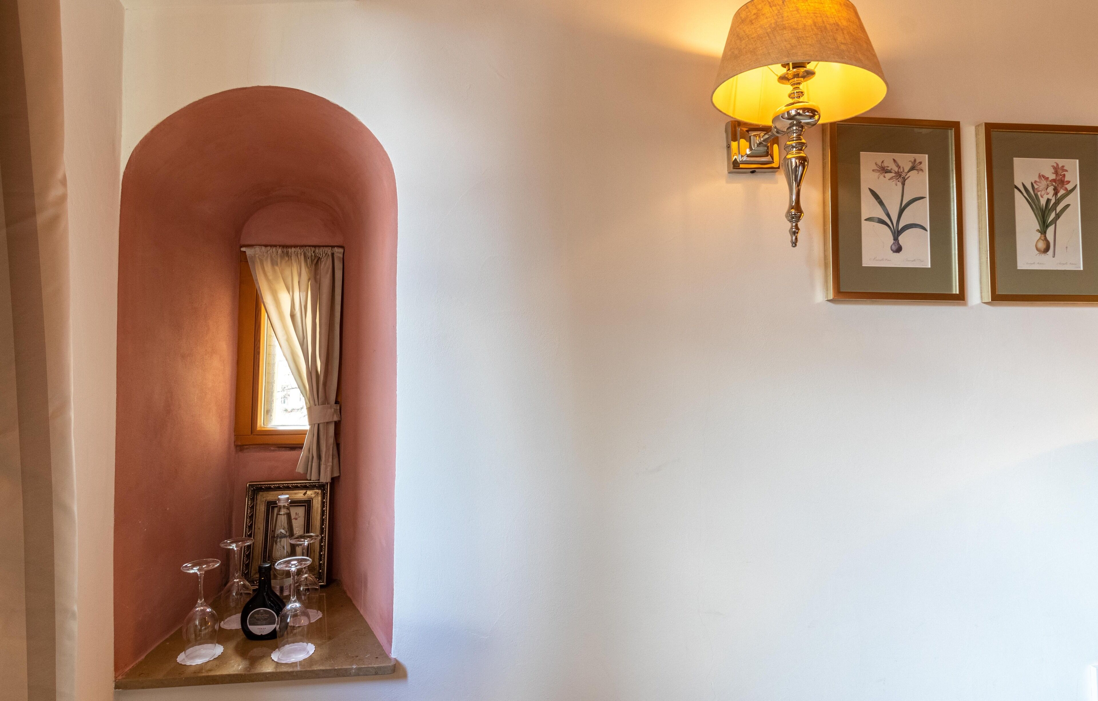 A room with a pink archway, a table with wine glasses and a bottle, a wall-mounted lamp, and two framed botanical illustrations.