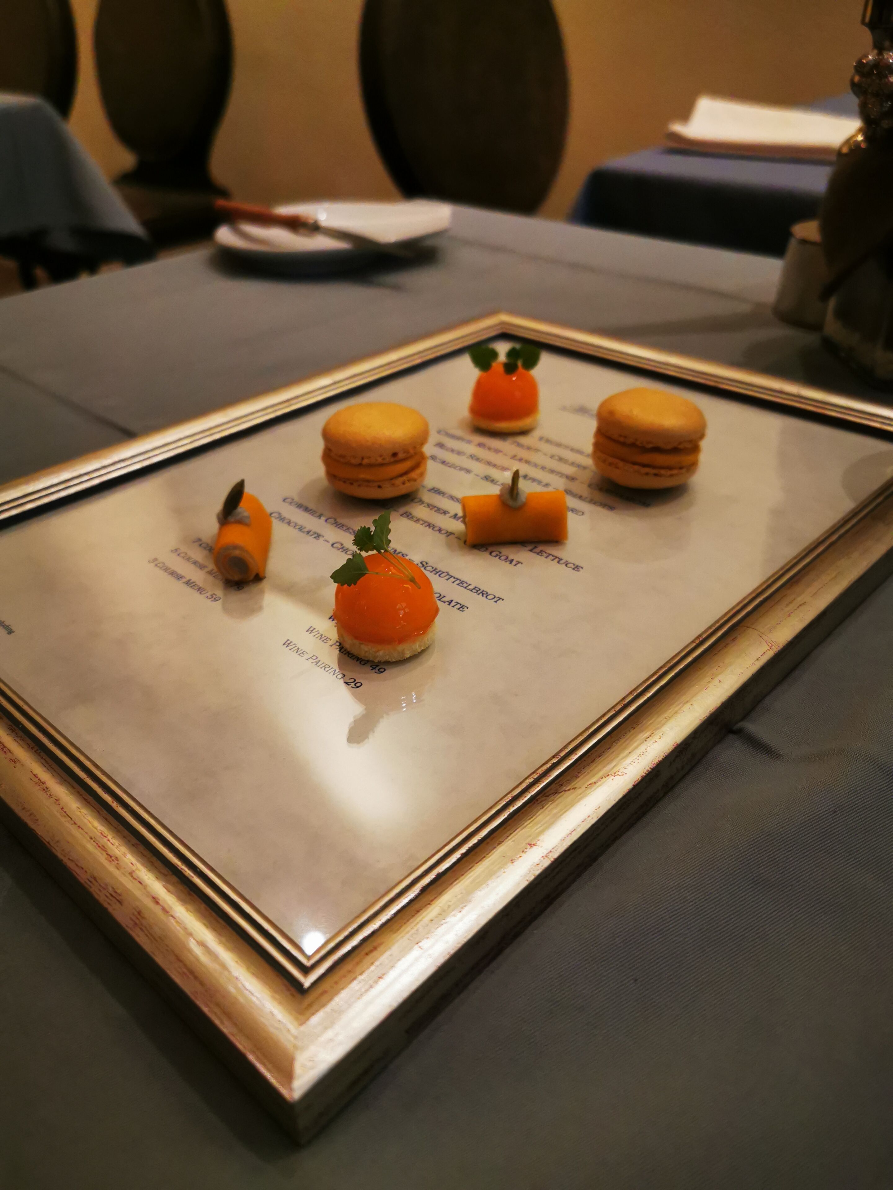 A framed menu with various food items, including macarons and a small orange garnish, displayed on a table.