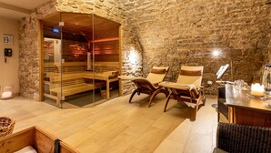 Un sauna con paredes de piedra, bancos y sillas de madera, puerta de vidrio y una mesa con velas y vasos.