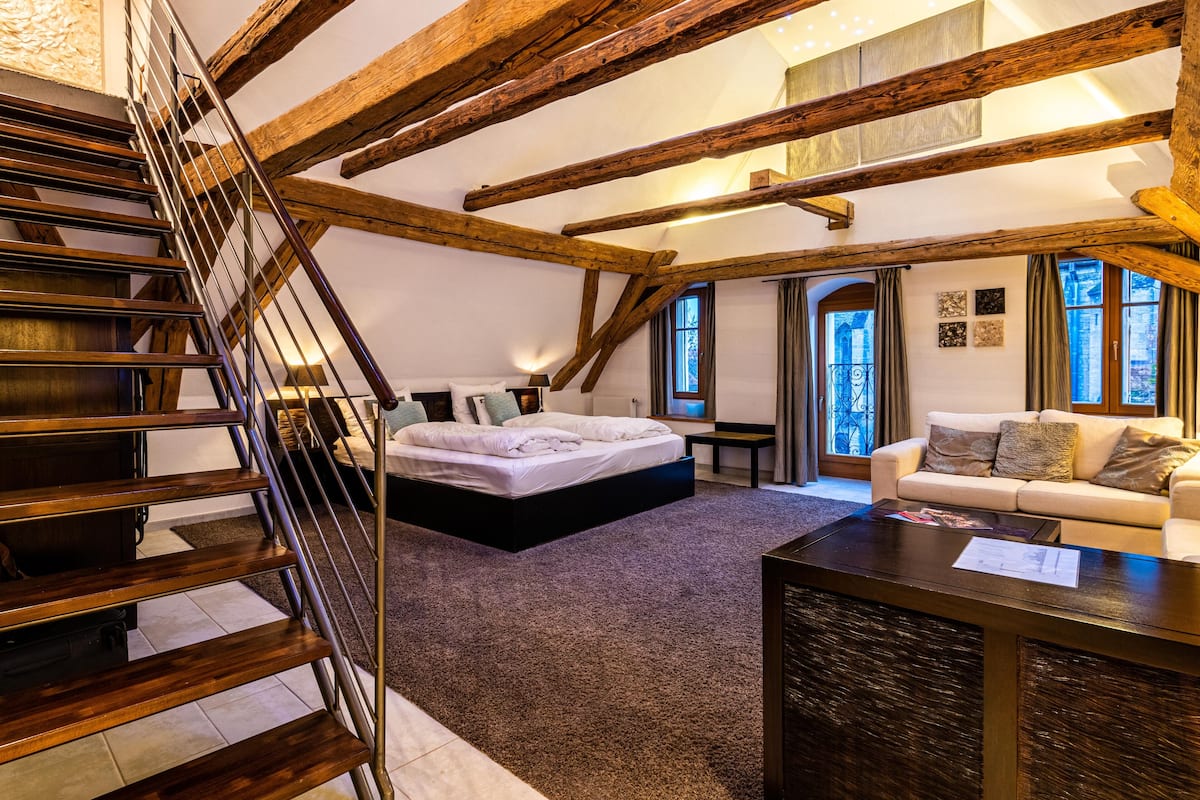 A bedroom with a large bed, a sofa, a coffee table, and a staircase with wooden beams.