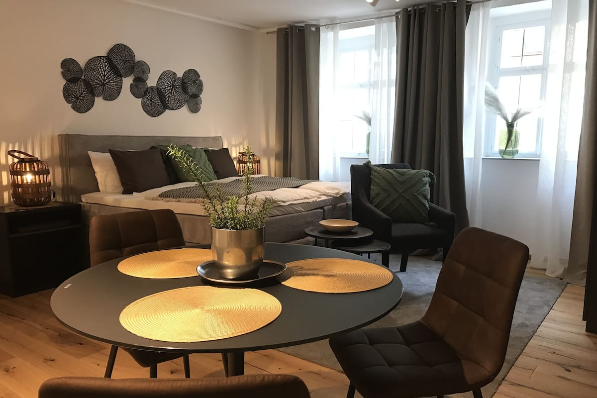 A modern bedroom with a bed, bedside tables, a nightstand, a potted plant, and a dining area with a table and chairs.