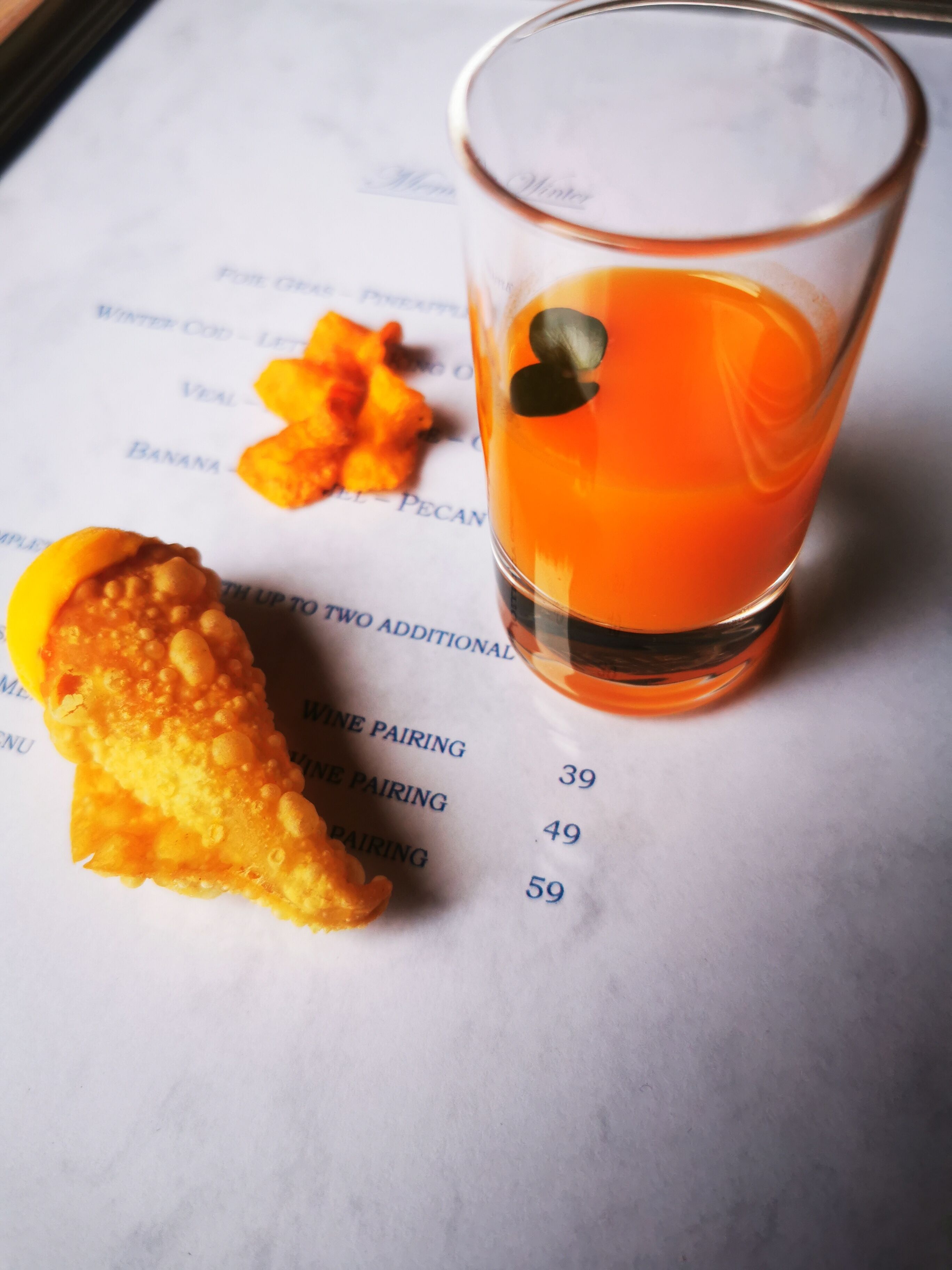 A menu with food items and prices, a glass of orange drink, and a piece of fried food.