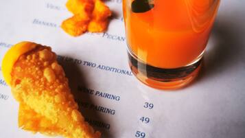 A menu with food items and prices, a glass of orange drink, and a piece of fried food.