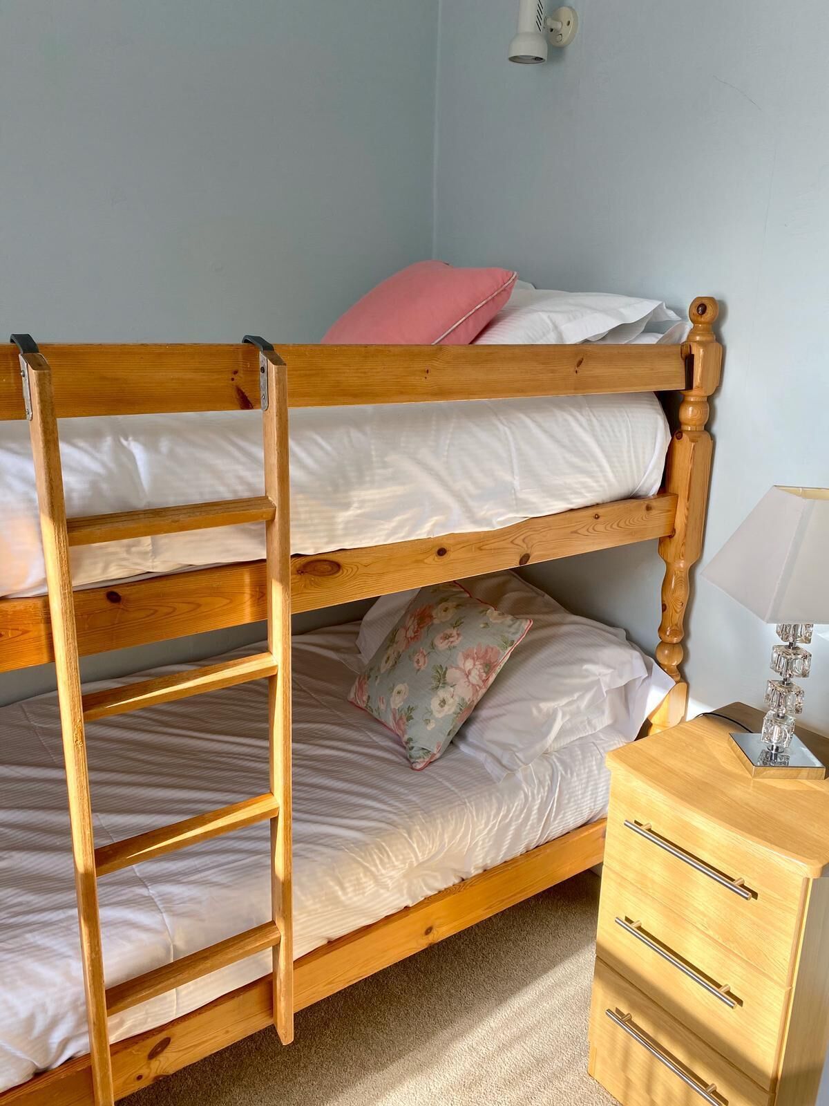 A bunk bed with a wooden ladder, a bedside table with drawers, and a lamp.
