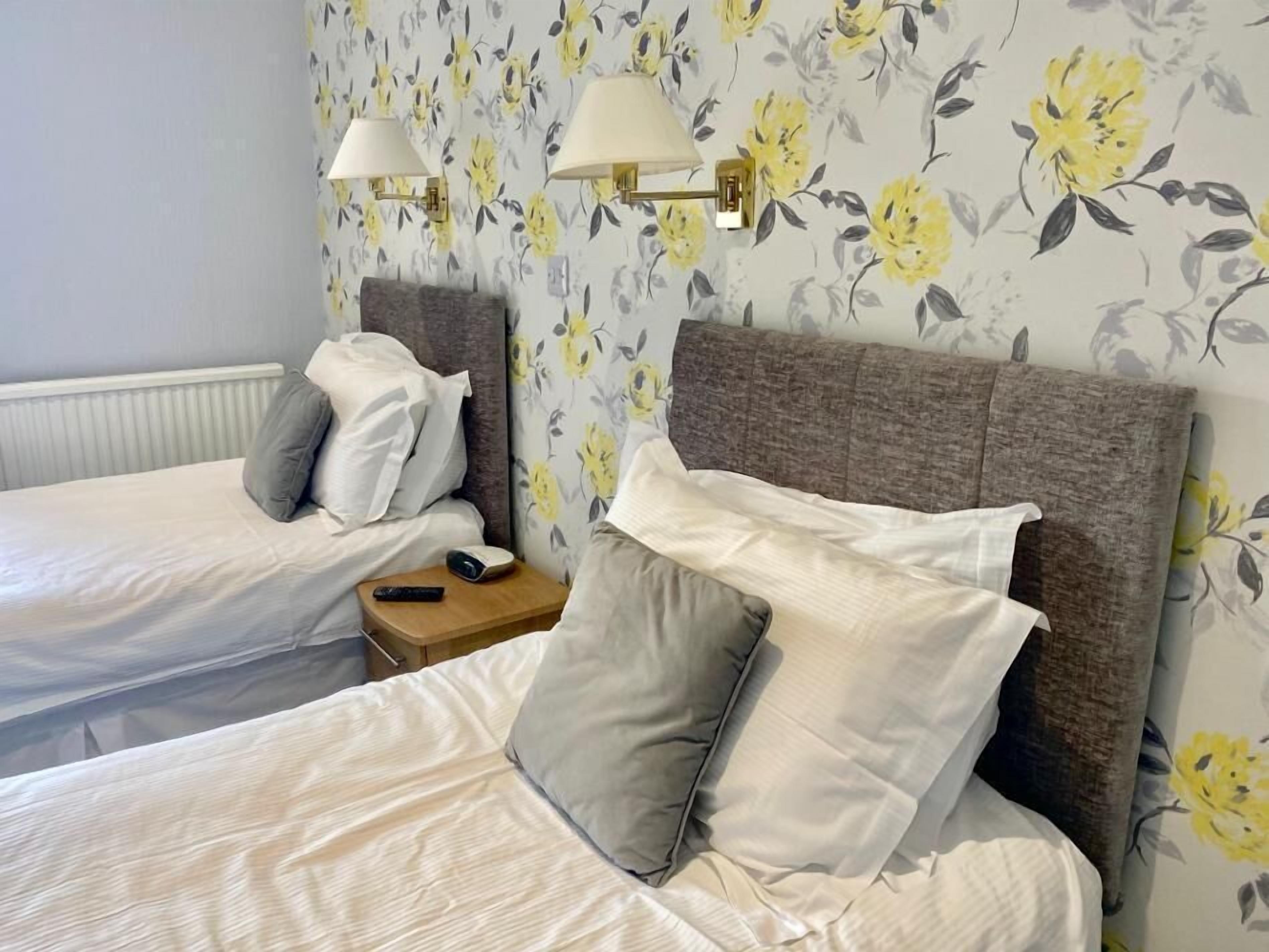 Ein Hotelzimmer mit zwei Betten, jeweils mit grauen Kopfteilen und weißer Bettwäsche. Zwei Nachttische mit Lampen und einer Fernbedienung auf einem. Die Wände sind mit einem floralen Wandpapier designt.