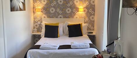 A neatly arranged hotel room with a bed, bedside lamps, a small table with a kettle and cups, and a trash bin.