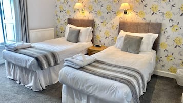 A hotel room with two beds, each with a headboard, bedside tables, and lamps. The walls have a floral wallpaper pattern.