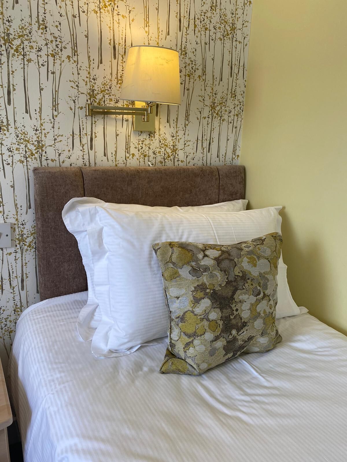 A neatly made bed with white and patterned pillows, a brown headboard, and a wall-mounted lamp.