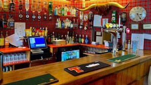 A bar with a red plaid wall, various liquor bottles, a clock, and a computer monitor. - The Kings Arms Hotel (Castle Douglas)