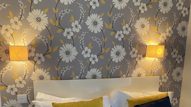 A neatly made bed with white and yellow pillows, a white headboard, and a floral wallpaper pattern. There are two bedside lamps on either side of the bed. - The Kings Arms Hotel (Castle Douglas)