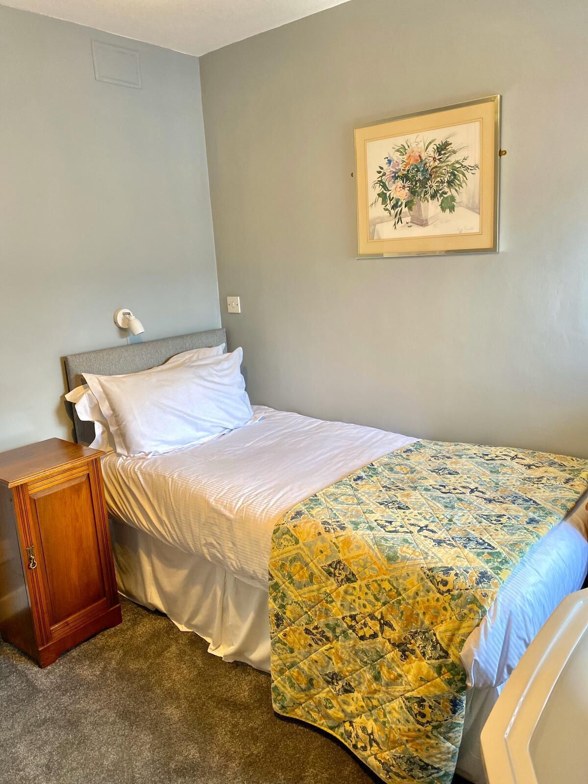 Un dormitorio con cama, mesita de noche, lámpara de pared y un cuadro floral enmarcado.