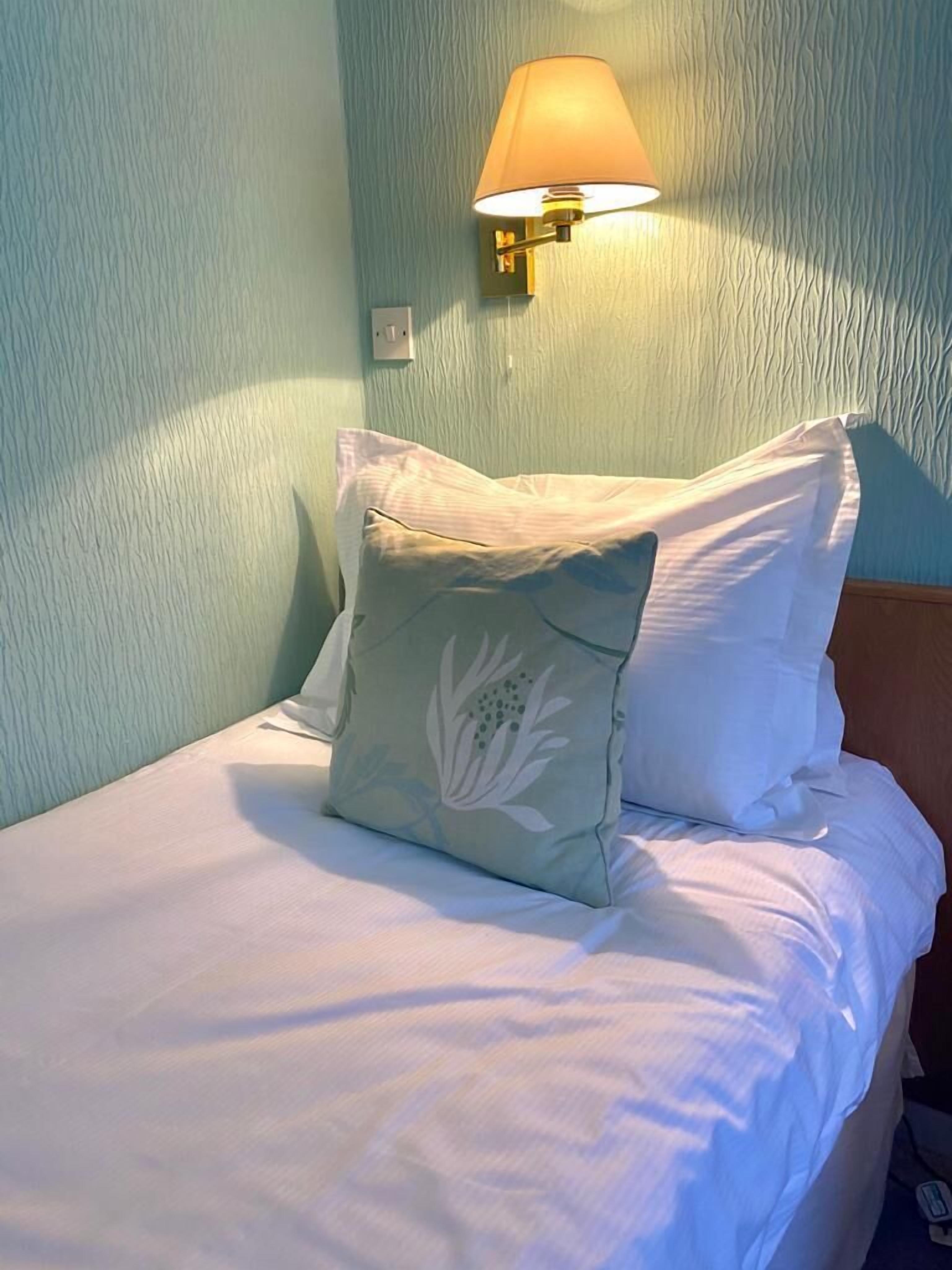 Un lit bien fait, avec du linge de lit blanc et deux oreillers, dont l’un est orné d’un motif floral, dans une pièce aux murs texturés et dotée d’une lampe fixée au mur.