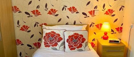 En snyggt bäddad säng med vita sängkläder och röda, blommiga kuddar, en sänglampa och ett träbord vid sängen med en digital klocka.