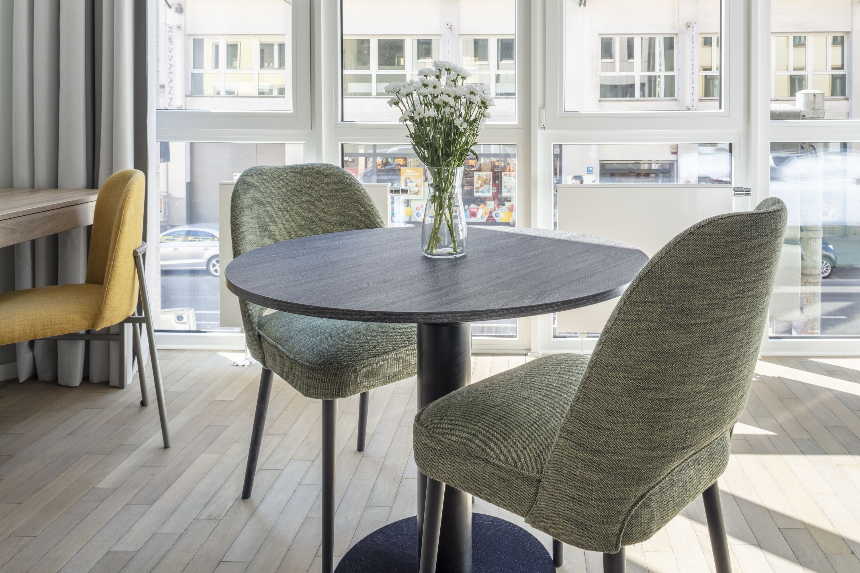 A modern room with a round table and two chairs, a vase with white flowers, and large windows offering a view of the street.
