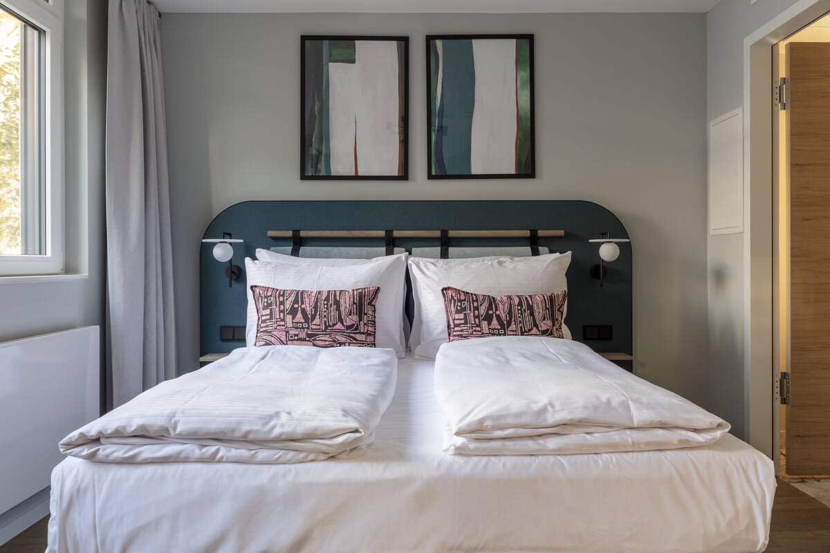 A double bed with white linens and patterned pillows, a dark headboard, and two abstract paintings on the wall.