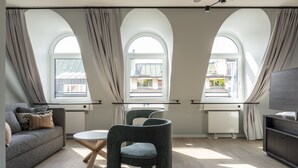 Ein modernes Wohnzimmer mit einem grauen Sofa, zwei Sesseln, einem Couchtisch und einem Fernseher auf einem Schrank. Drei Bogenfenster mit Vorhängen und ein Heizkörper an der Wand sind zu sehen.