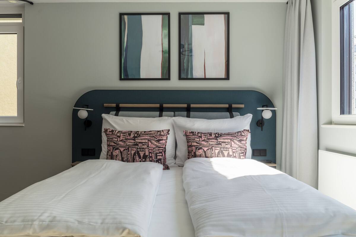 A double bed with patterned pillows, a headboard with wall-mounted lights, and two framed artworks above.