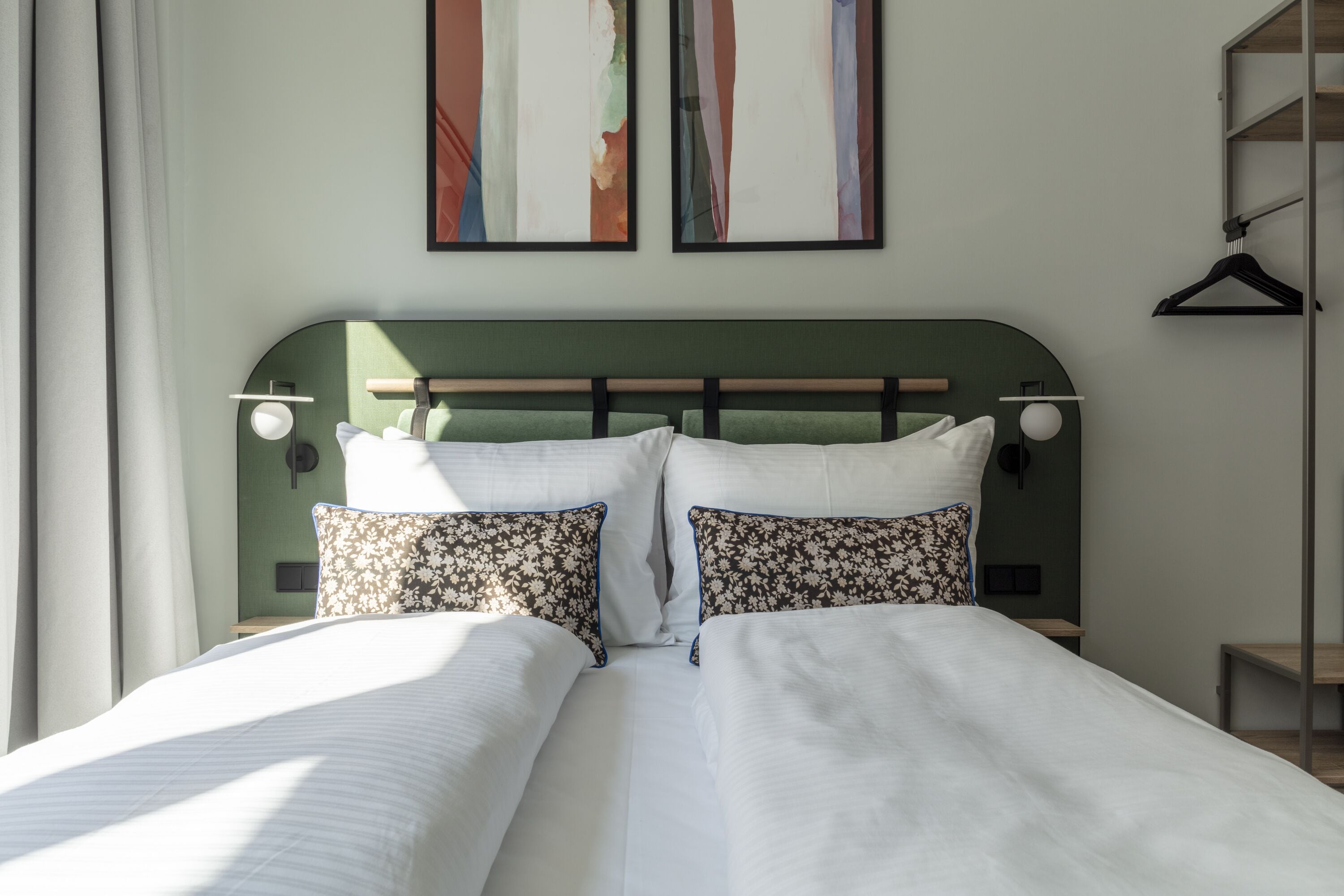A neatly made bed with white linens and floral pillows, a green headboard, and two wall-mounted lamps.