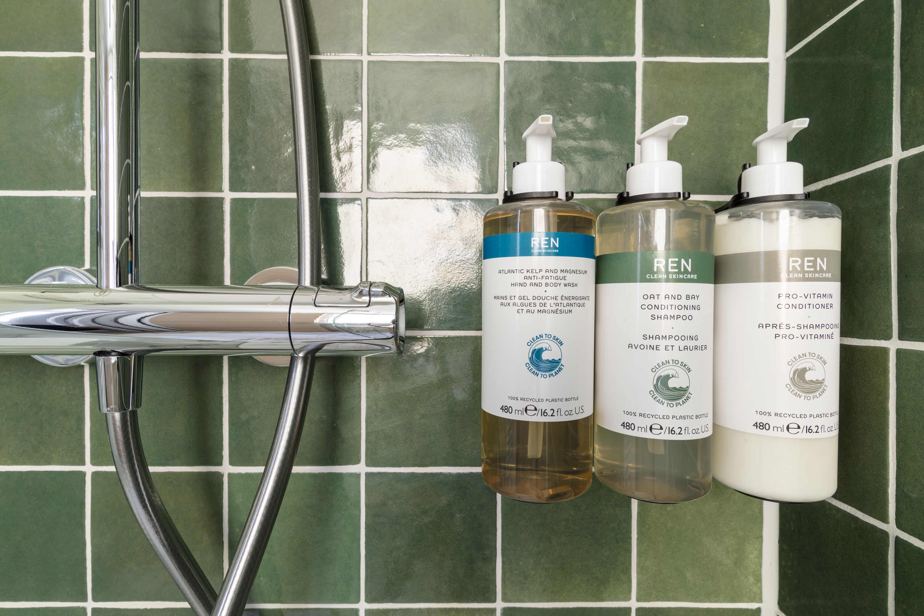 Três frascos de produtos de tratamento de pele REN apoiados em uma parede revestida de azulejos.