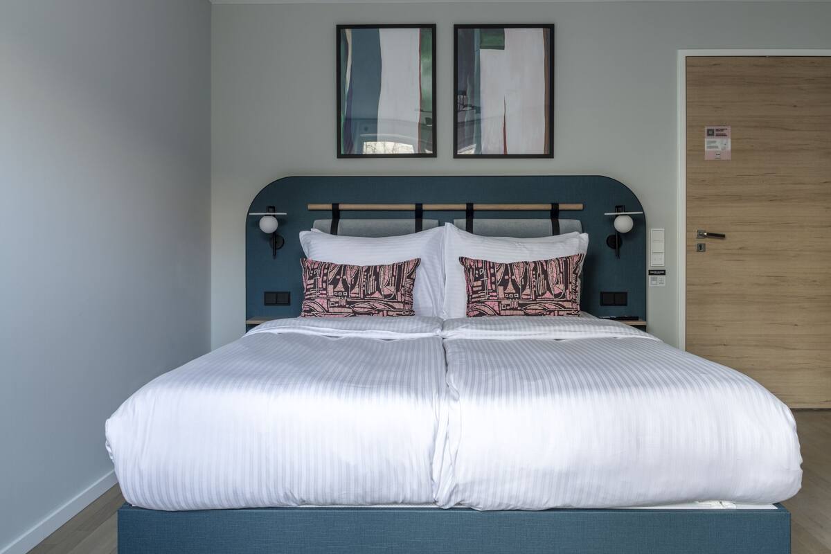 A neatly made bed with white bedding and patterned pillows, a blue headboard, and two framed artworks above it.