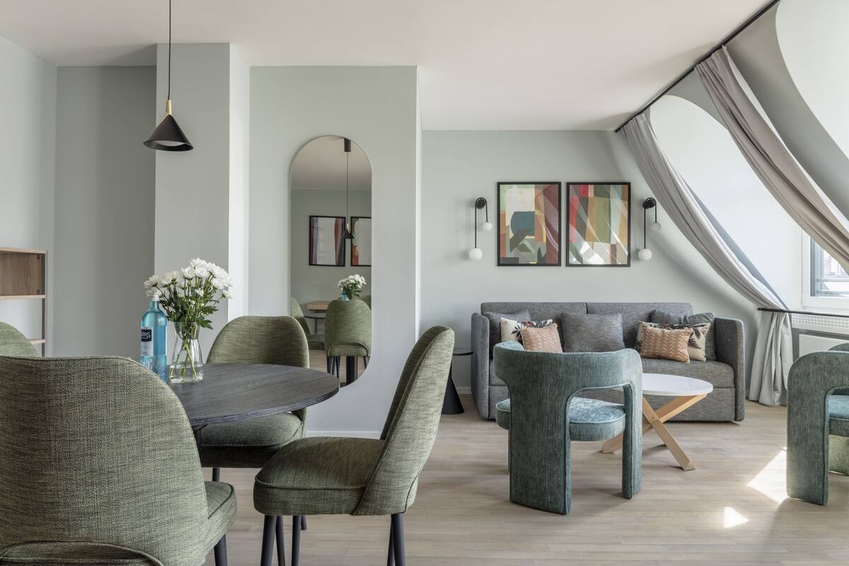 A modern living room with a dining area, grey sofas, and a large window with curtains.
