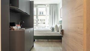 Ein kleines Hotelzimmer mit einer Küchenzeile, einem Bett mit Kissen, einem Fenster mit Vorhängen und einer Holzwand.